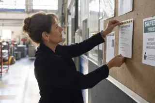 Woman hanging a safety checklist on a bulletin board in an industrial corridor with soft natural light, calm and focused atmosphere.
