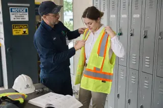 Technician in industrial workspace pausing with safety gear near open electrical panel, glancing at a checklist, sunlight streaming in, calm focused atmosphere
